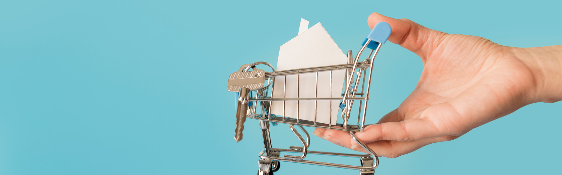 close-up-hand-holding-miniature-shopping-cart-with-paper-house-keys-against-blue-background.jpg